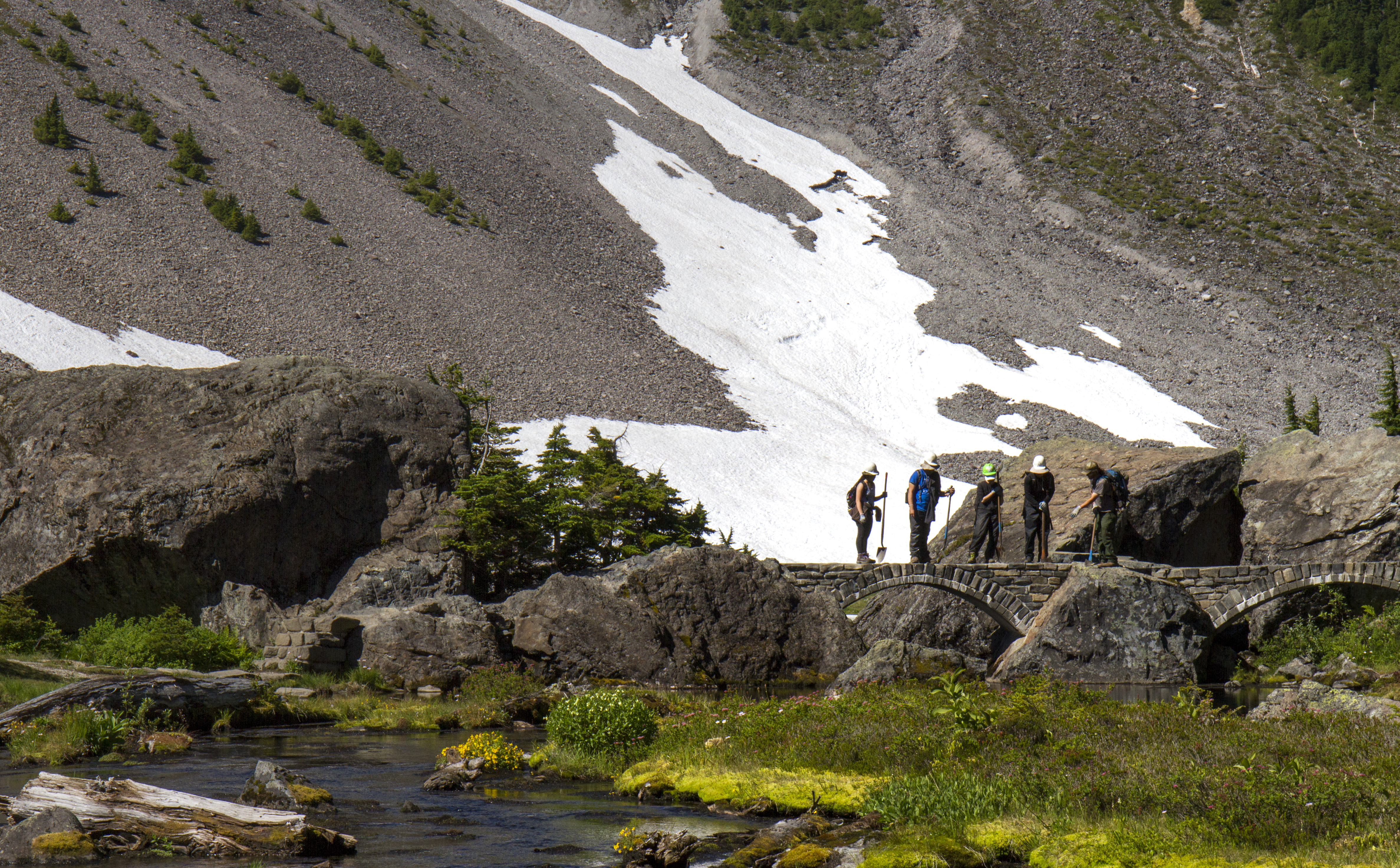 long shot of five people standing on a bridge above a stream holding shovels. behind them is a sloping shale covered hill with a patch of snow. the stream is surrounded by bus sized boulders